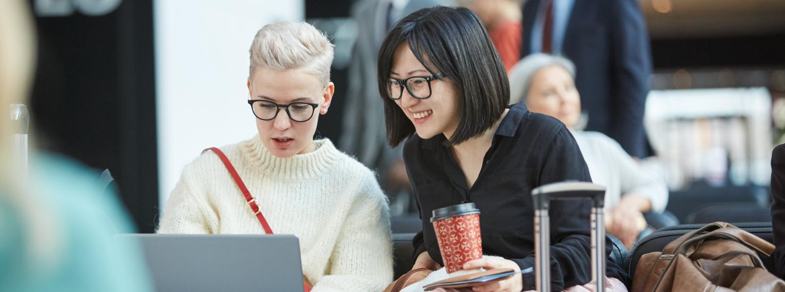 Zwei Studentinnen mit Gepäck und Laptop am warten an einem Flughafen.