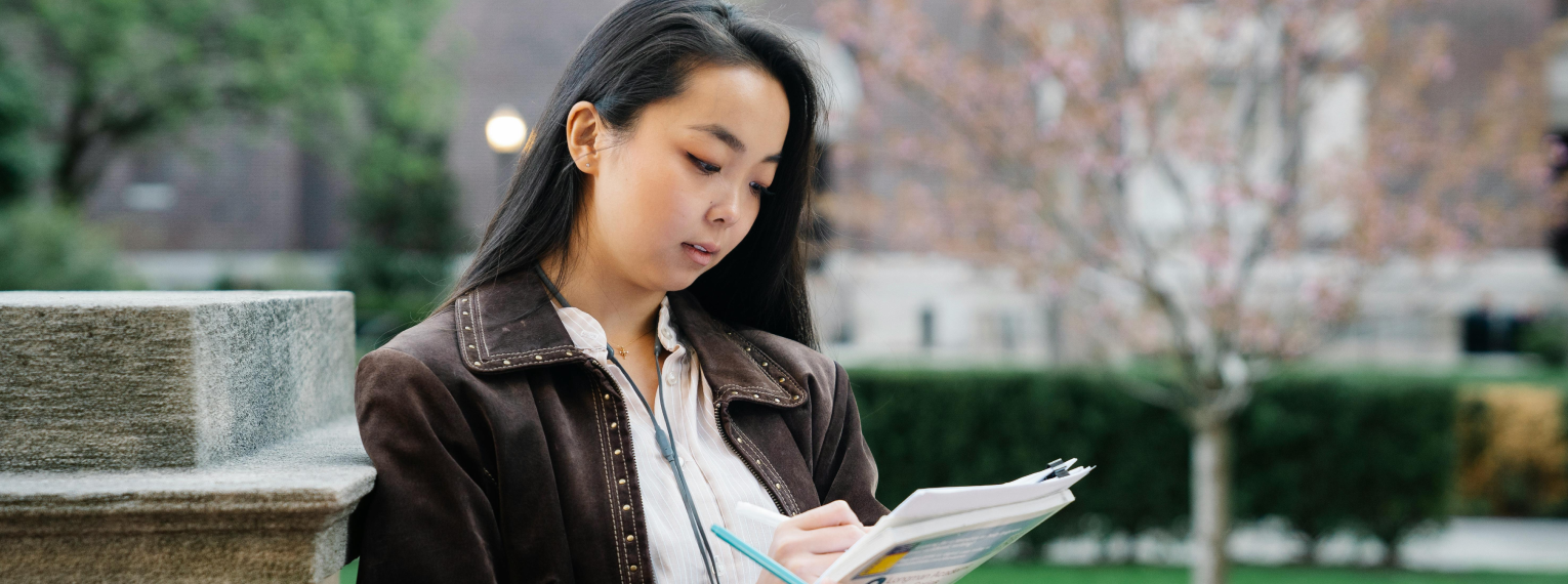 Eine Studentin sitzt draußen auf dem Campus und macht sich Notizen.