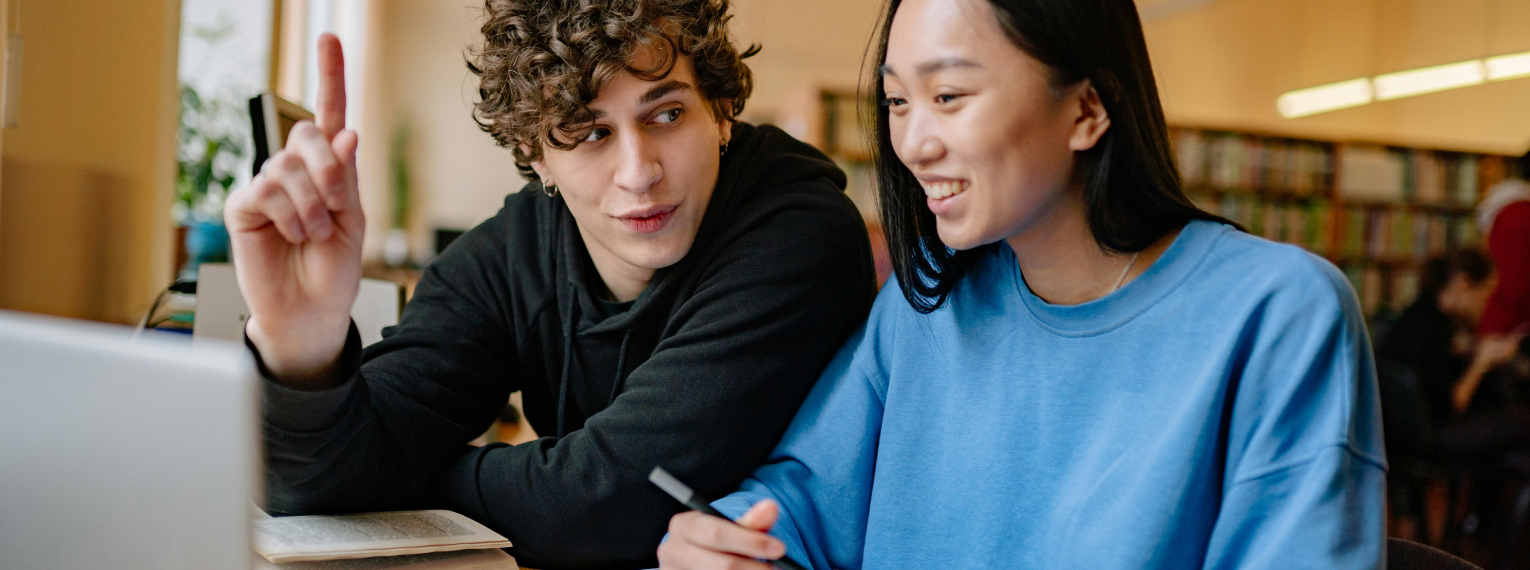 Zwei Studierende sitzen vor einem Laptop in einer Bibliothek.