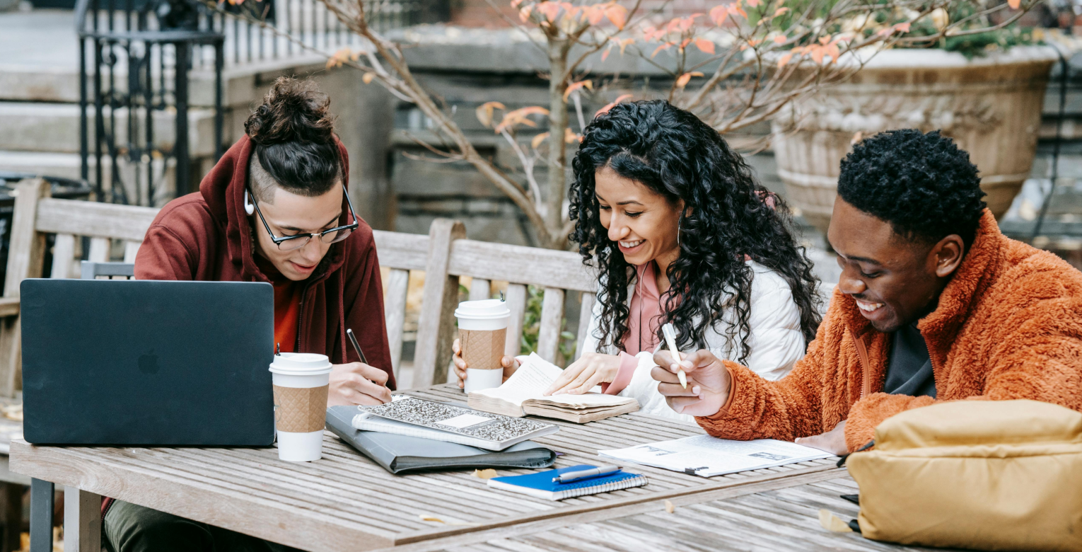 Drei Studierende sitzen arbeitend an einem Tisch im Freien. 